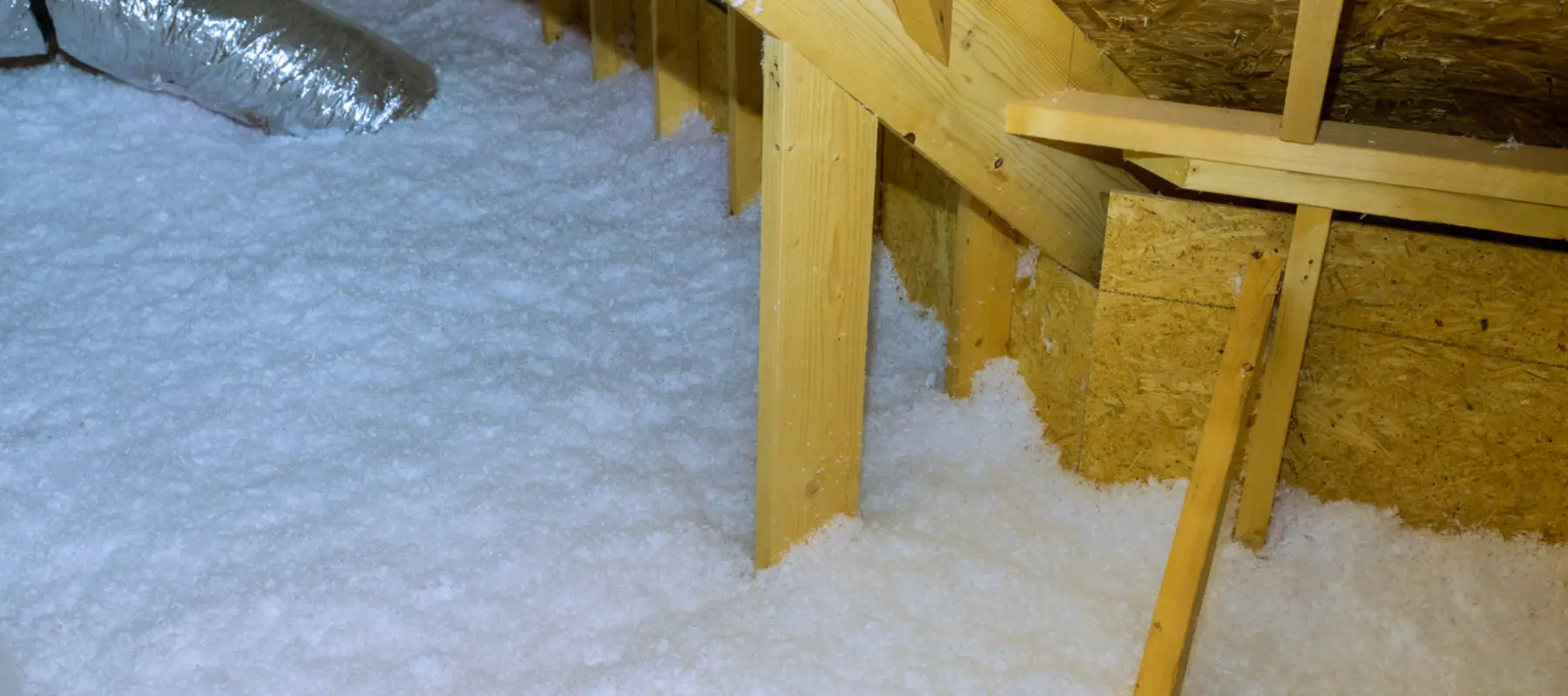 Attic with wooden beams and blown-in white cellulose insulation covering the floor. A silver ventilation duct is visible in the upper left corner.