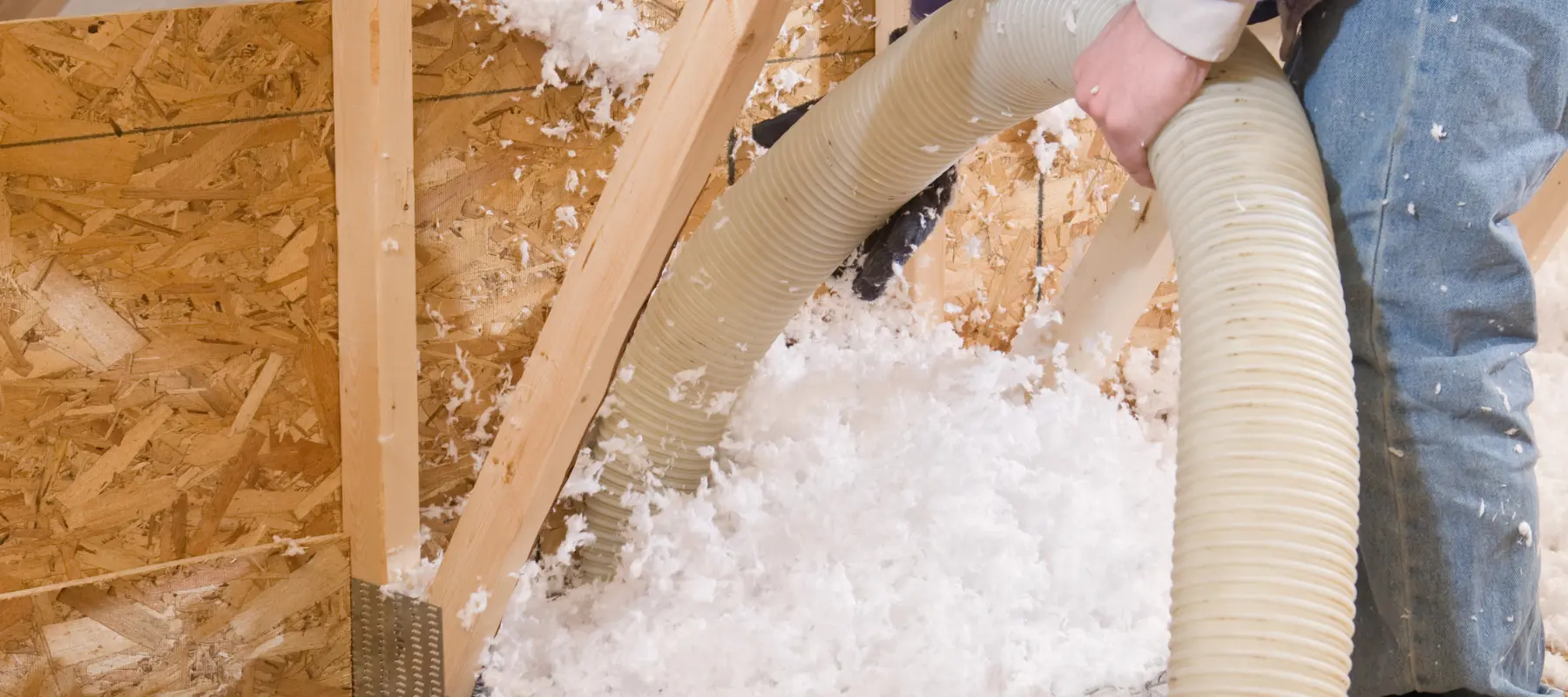 worker spraying brown fiberglass insulation between attic trusses