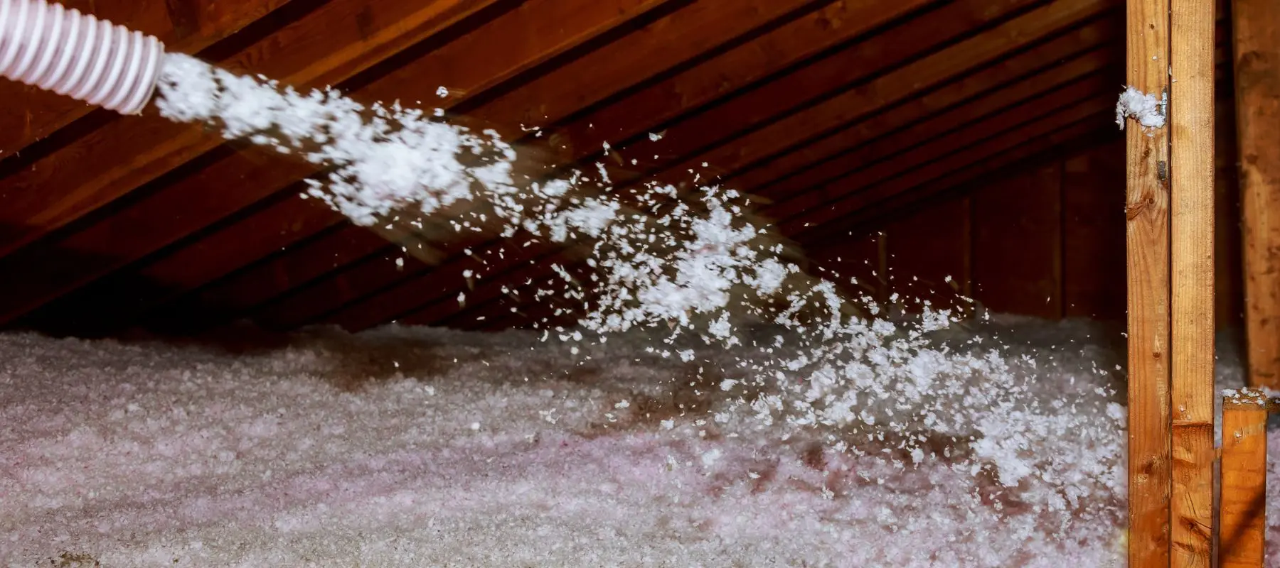 A hose sprays loose fill insulation into an attic with exposed wooden beams, creating a fluffy white layer on the floor.