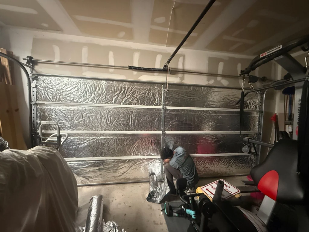 A person kneels on a garage floor, installing reflective insulation panels on a closed garage door. Gym equipment and exercise items are visible nearby.
