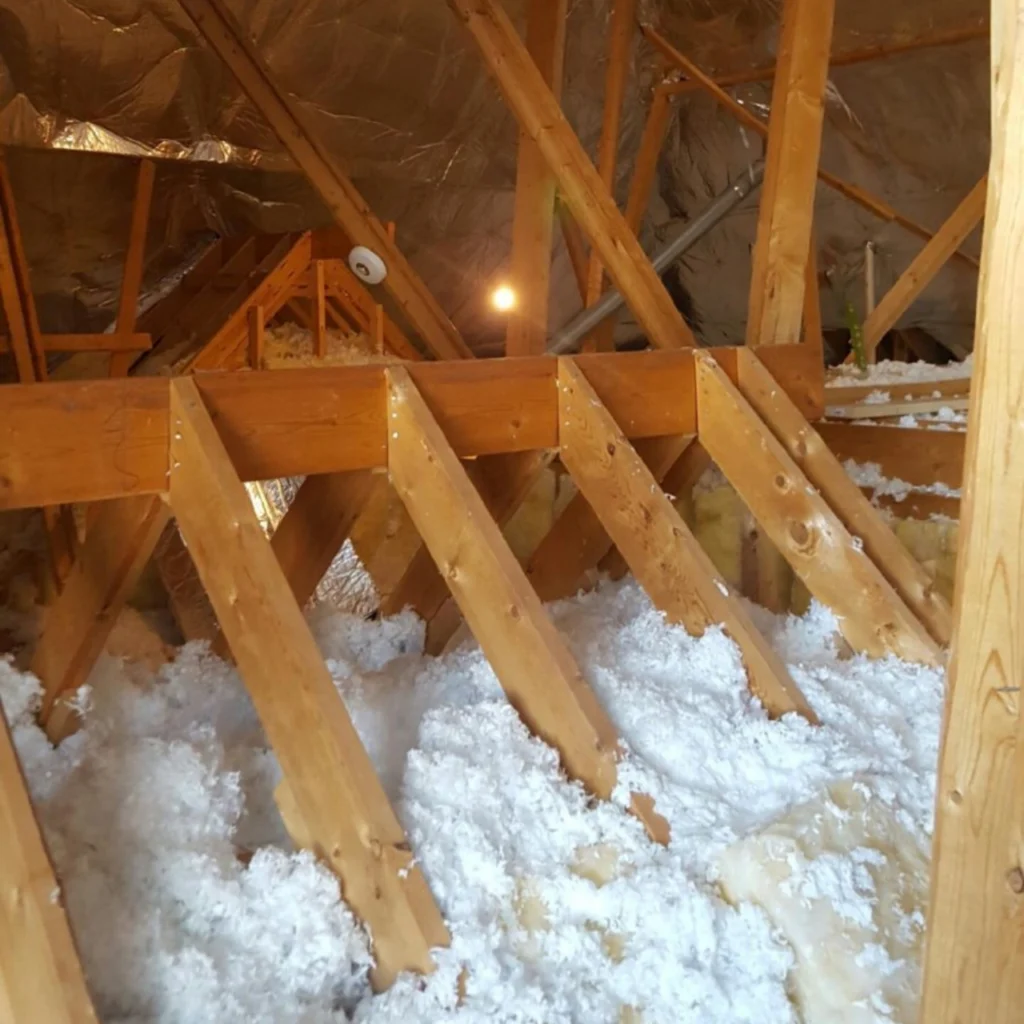 Wooden beams and rafters in an attic with loose-fill insulation covering the floor. Light shines in the background, and reflective insulation is attached to the roof.