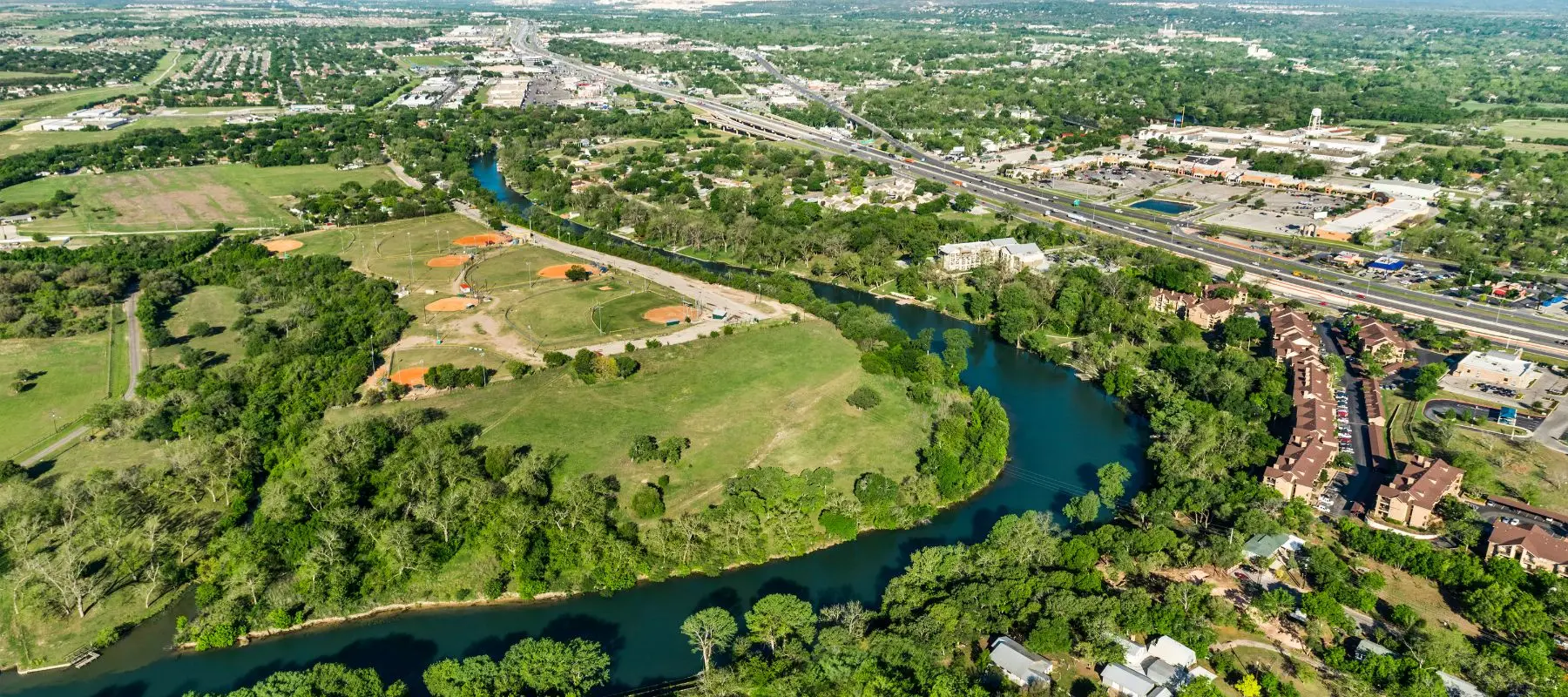 Aerial view of a winding river surrounded by green trees, a baseball field complex, nearby roads, buildings, and a highway stretching into the distance under a clear sky.