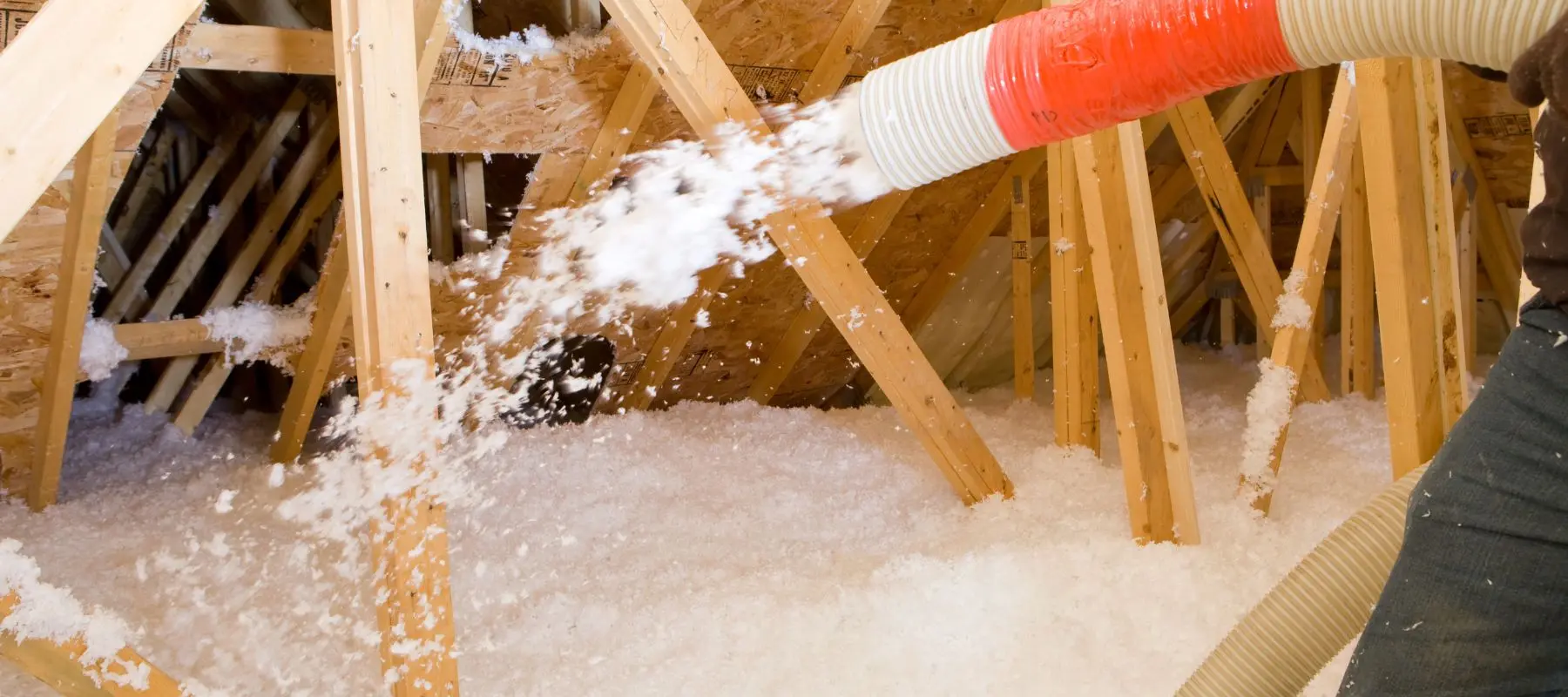 A person uses a large hose to blow loose-fill insulation into an unfinished attic, covering the floor between exposed wooden beams.