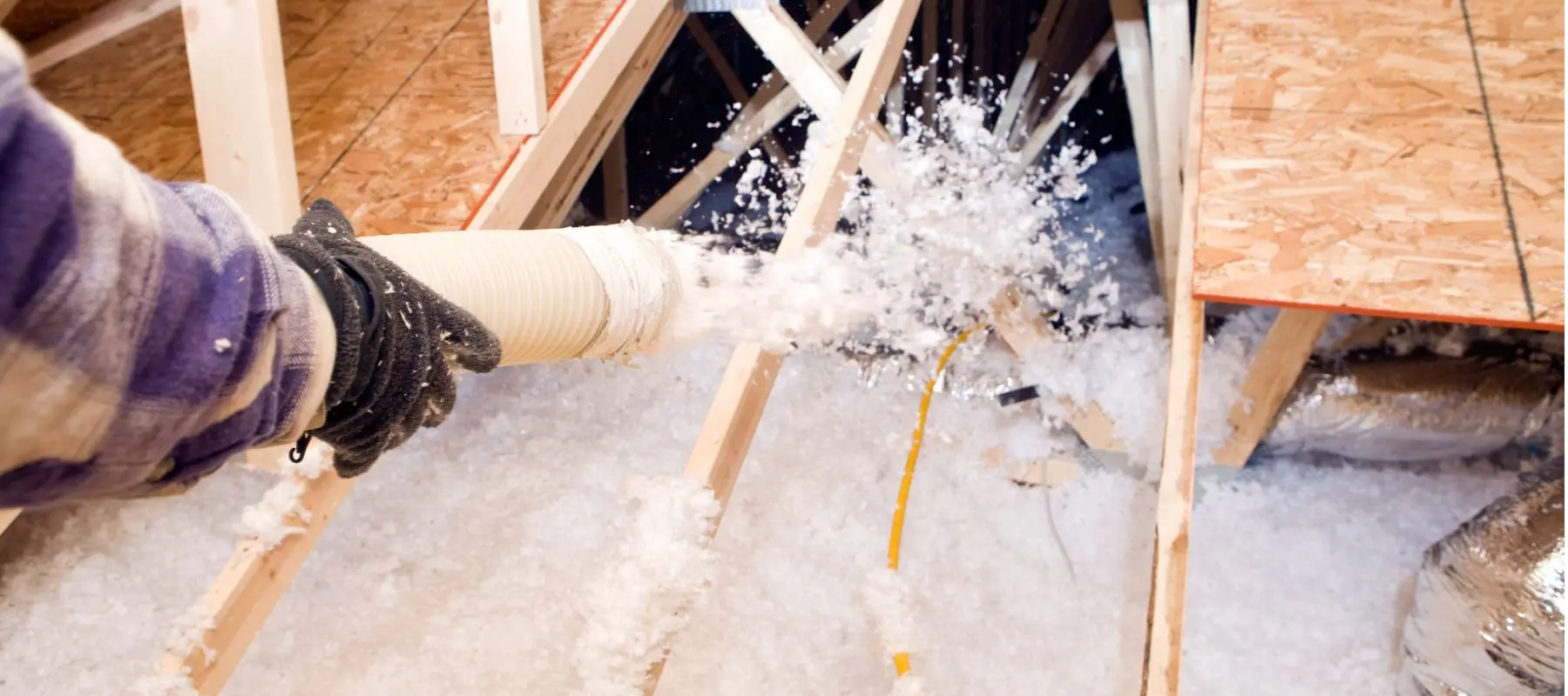 A gloved hand sprays loose-fill insulation into an attic, covering wooden beams and electrical wiring with white insulating material.