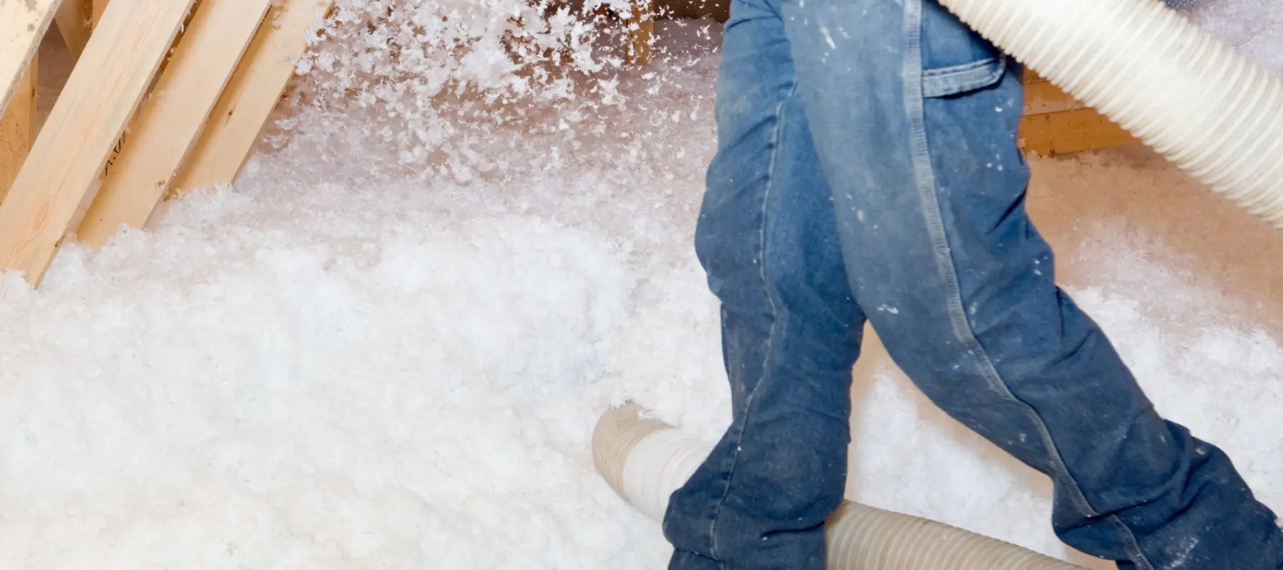 Person in blue jeans blowing white insulation material into an attic, with insulation spraying out from a large hose.