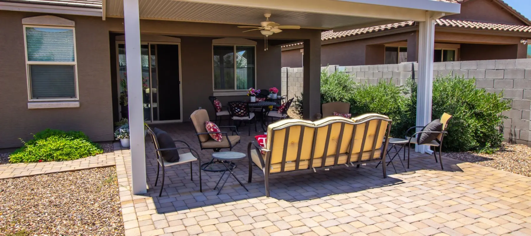 Covered patio with cushioned seating, round table, chairs, ceiling fan, and paver floor. Surrounded by plants, gravel, and a block wall, adjacent to a house with windows and sliding glass doors.