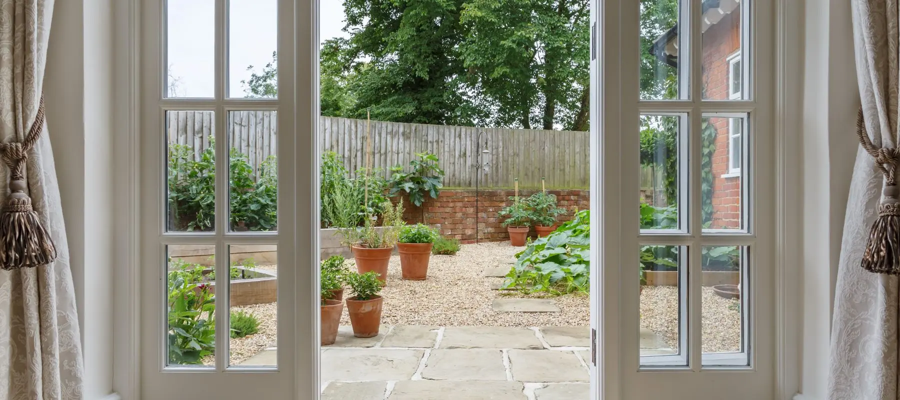 View through glass French doors to a garden with potted plants, raised beds, gravel paths, and a wooden fence, with trees and part of a brick house visible.