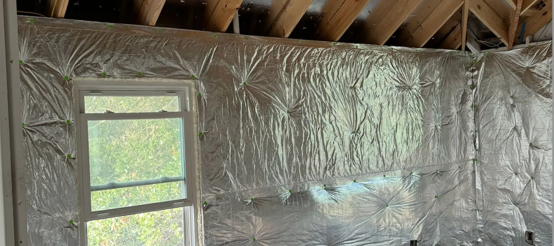 A room with exposed beams and walls covered in reflective insulation material. A window lets in natural light and shows greenery outside.