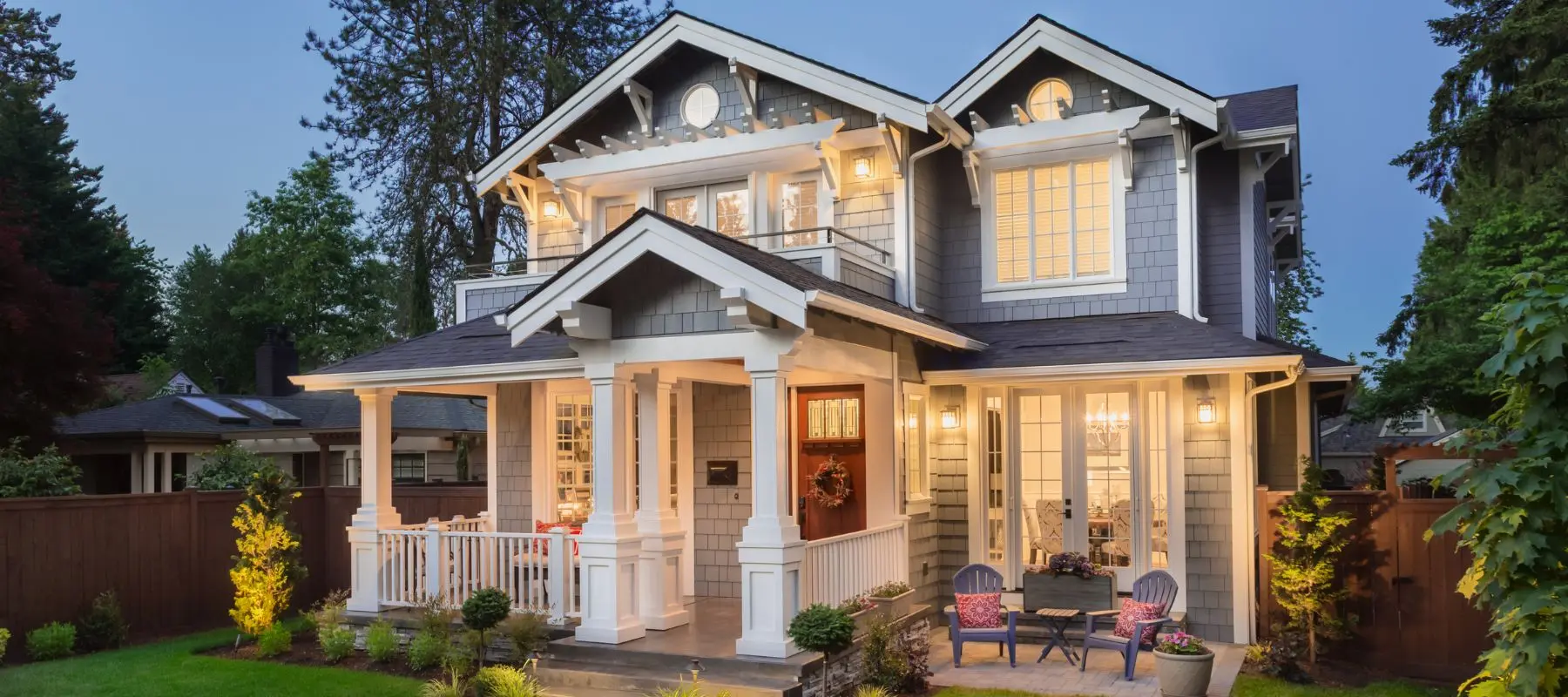 Two-story gray house with white trim, large windows, and a front porch. The yard has outdoor seating, plants, and warm lighting, creating a welcoming evening atmosphere.
