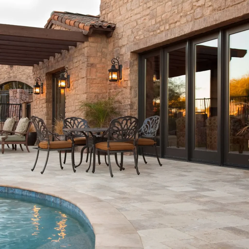 Elegant stone patio with wrought iron table and chairs beside a curved swimming pool. Large glass doors and lantern-style wall lights decorate the stone exterior of the house. Potted plants add greenery to the scene.
