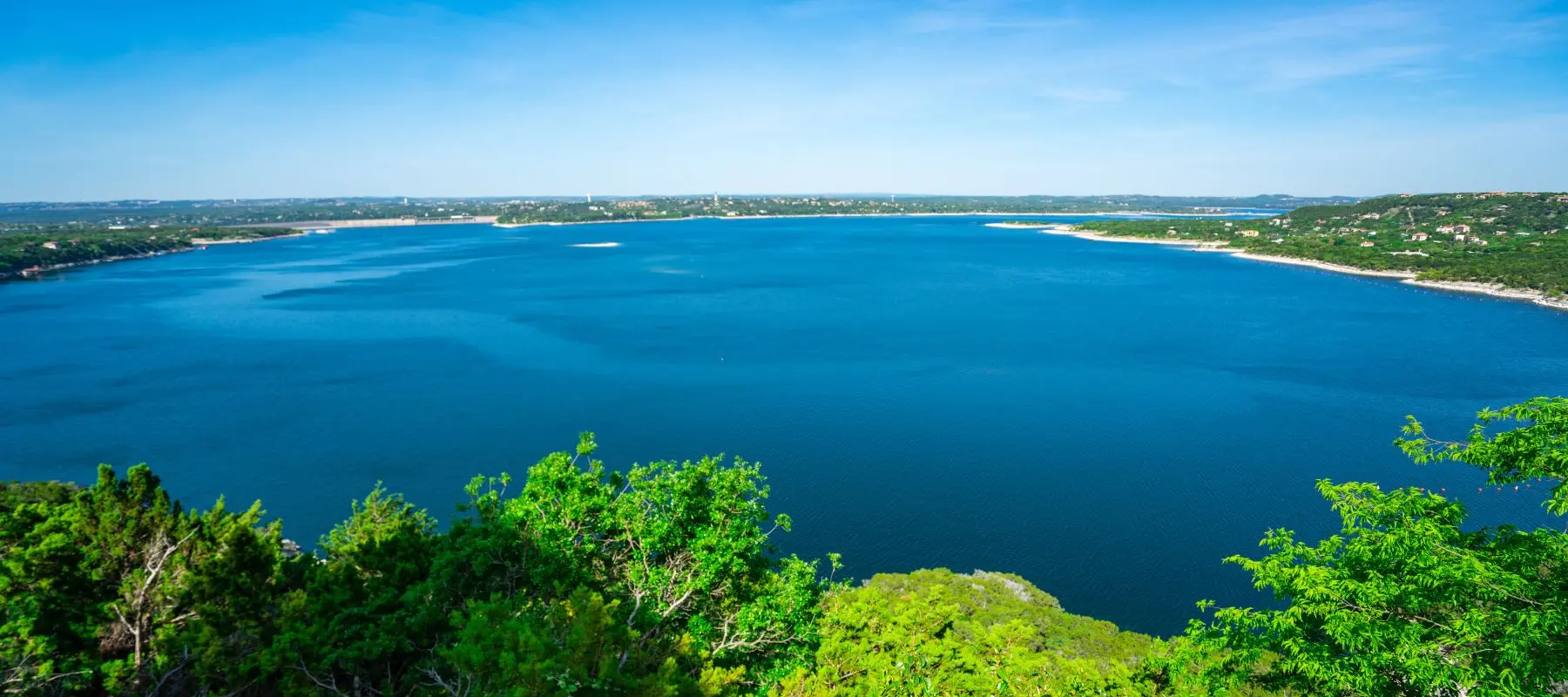 A wide, deep blue lake surrounded by lush green trees and rolling hills under a clear, bright blue sky.
