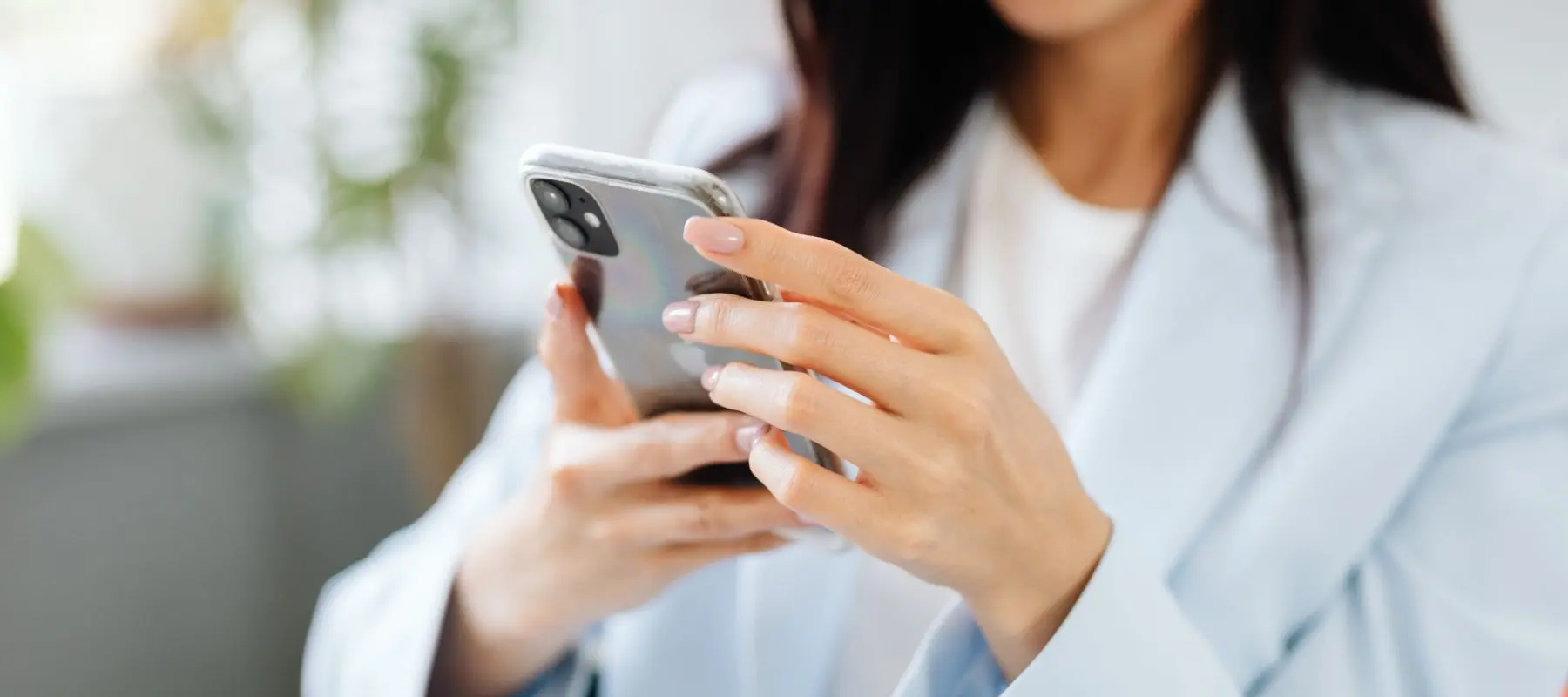 A woman wearing a light blue blazer holds and uses a smartphone, focusing on the device. Her face is partially out of frame, and the background is softly blurred.