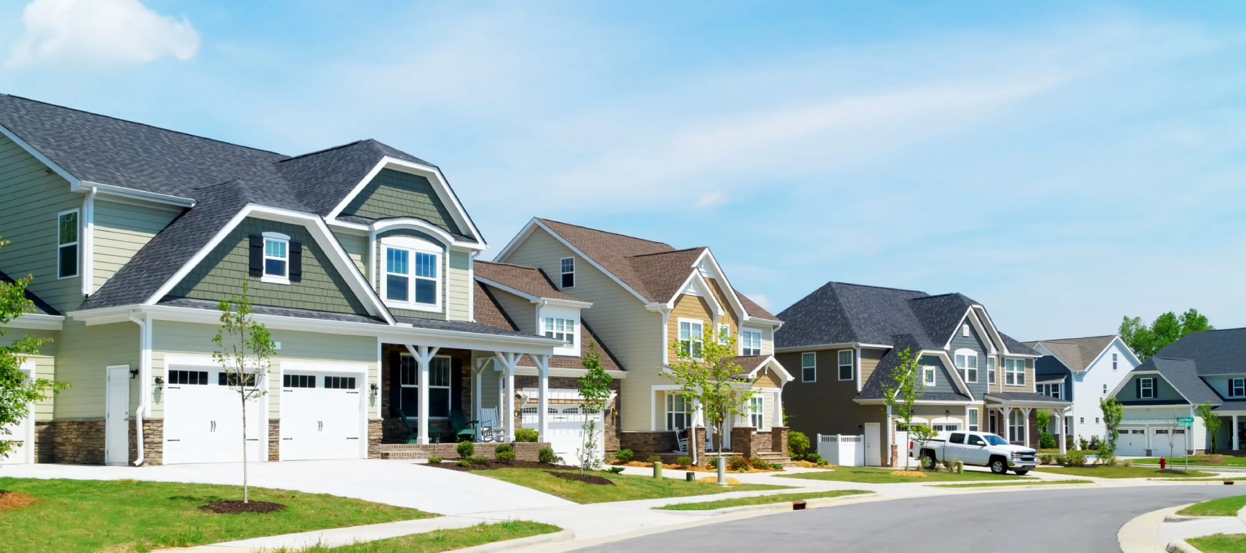 A row of modern, suburban houses with garages and well-kept lawns lines a quiet, curved street under a blue sky with light clouds.