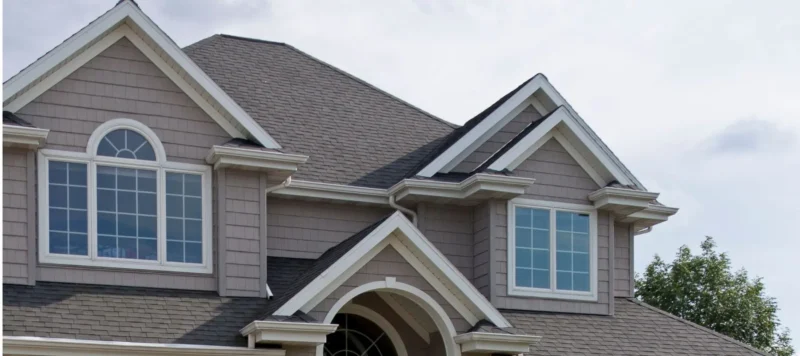 The upper portion of a house with gray siding, white trim, large arched and rectangular windows, and multiple peaked gables against a cloudy sky.