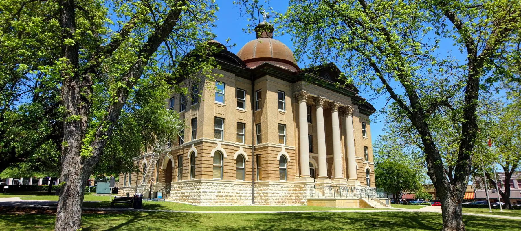Historic courthouse with a large dome, tall columns, and arched windows, surrounded by green trees and grass on a sunny day.